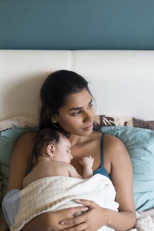 Young mother lying in bed with her baby son asleep on her chest. They both have a blanket over them and the mother is looking to the side.の写真素材