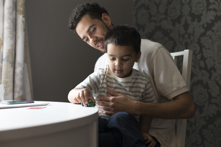 Little boy is sitting on his father's knee, playing with a toy car on the table.の写真素材