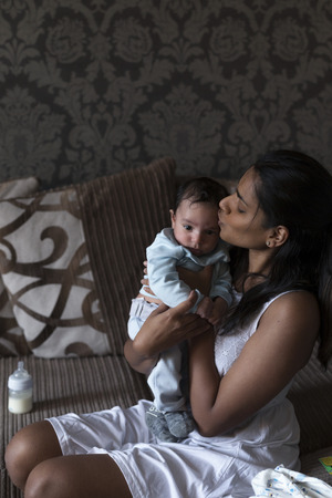 Young mother is sitting on her sofa and holding her baby son who has just been fed. She is kissing him on the head with him held close to her.の写真素材