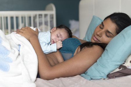 Young mother having a nap in bed with her baby son sleeping on her chest.の写真素材