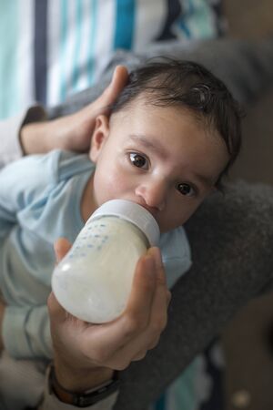 Young father is sitting with his baby son in his lap. He is feeding him a bottle of milk.の写真素材