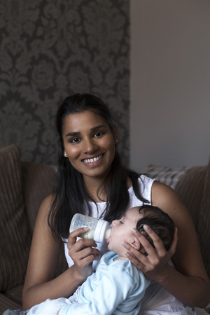 Young mother is smiling for the camera as she sits and feeds her baby son a bottle of milk.の写真素材