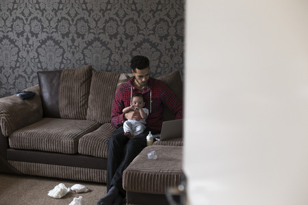 Young father sitting on his sofa at home with his baby son on his knee. He is working on a laptop and there are dirty nappies on the floor.の写真素材