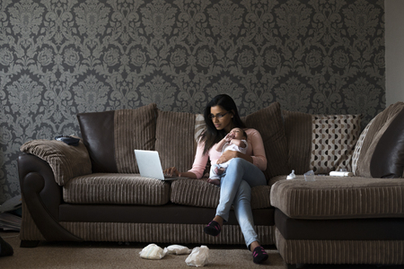 Young mother sitting on her sofa at home with her baby son on her knee. She is working on a laptop and there are dirty nappies on the floor.の写真素材