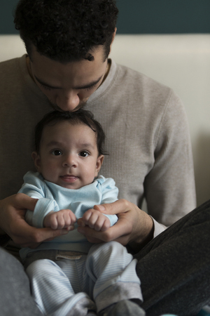 Young father sitting with his baby son in his lap. He is holding his hands and kissing him on his head. The baby is looking at the camera.の写真素材