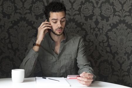Young man sitting at the table at home, using his smartphone and his bank card to pay bills over the phone.の写真素材