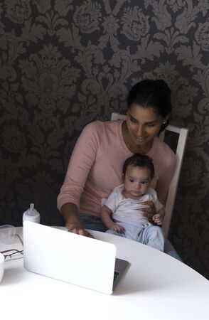 Young mother sitting at the table with her baby son on her lap, using a laptop.の写真素材