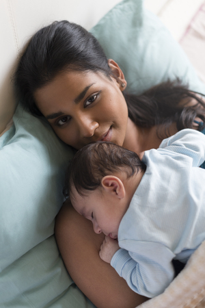 Young mother lying in bed with her baby son sleeping on her chest. She is smiling at the camera.の写真素材