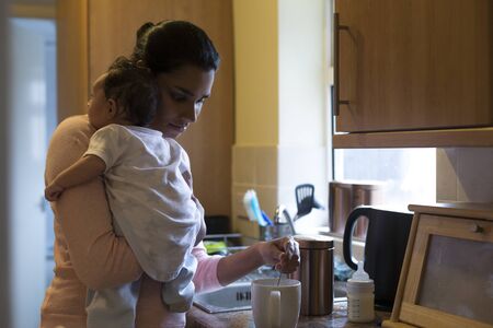 Young mother making a cup of tea with her baby son on her shoulderの写真素材
