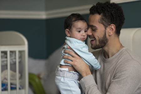 Young father sitting on his bed, holding his baby son. He has his nose pressed to his son's cheek.