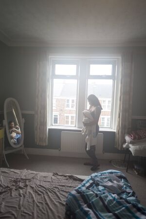 Young mother standing in her bedroom near the window with her sleeping baby son in her arms.の写真素材