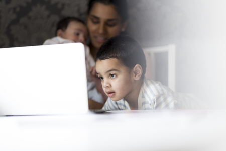 Little boy watching cartoons on his mothers laptop while she feeds her baby son in the backgroundの写真素材
