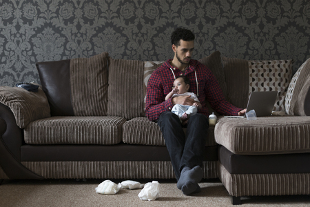 Young father sitting on his sofa at home with his baby son on his knee. He is working on a laptop and there are dirty nappies on the floor.の写真素材