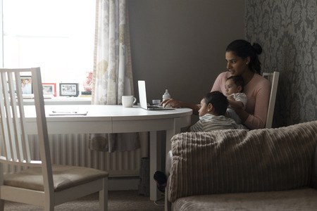 Young mother keeping her children entertained by showing them something on her laptop. She is holding her baby son and the eldest is leaning on her leg whilst looking at the screen.の写真素材
