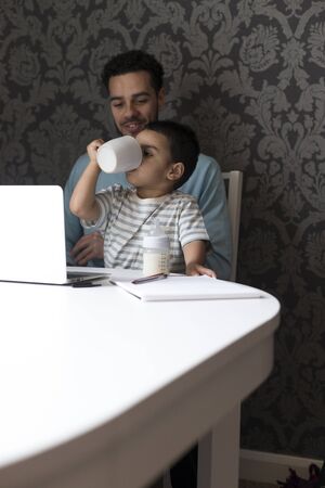 Young father is sitting at the table with his son on his lap. His son is copying what he has seen his dad doing and has his fathers mug to his mouth, pretending to drink his coffee whilst sitting at his computer .の写真素材