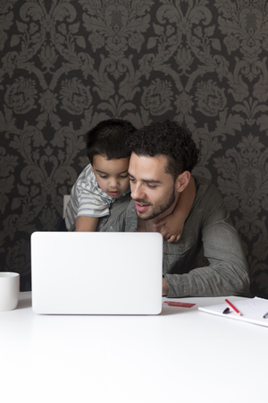 Little boy is climbing on his dad, trying to help him with his internet banking. They are sitting behind a laptop at home.の写真素材