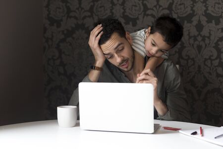 Little boy is climbing on his dad, trying to help him with his internet banking. They are sitting behind a laptop at home.の写真素材