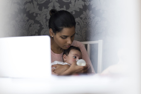 Young mother feeding her baby son a bottle of milk whilst sitting at her computer.の写真素材