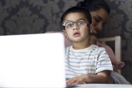 Little boy is imitating his mother by attempting to put her glasses on and typing on the laptop. His mother is in the background, feeding her baby son.の写真素材