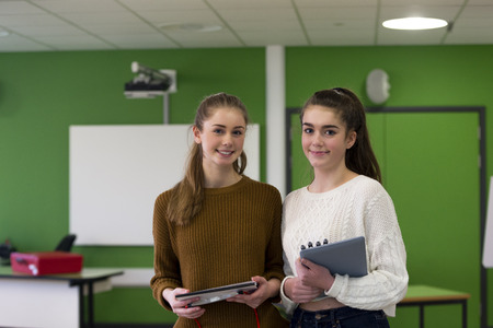 Two teenage girls standing together in a classroom with school eqipment in their hands.の写真素材