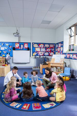 Group of nursery children sitting on the floor in their classroom with their teachers. The female teacher is reading from a book.の写真素材
