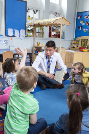 children are being read a story in nursery by a male teacher. One child has his hand up and the teacher is talking to him.の写真素材