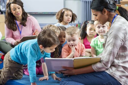 Female teacher giving a lesson to nursery students. They are sitting on the floor and there is a teacher taking notes.の写真素材