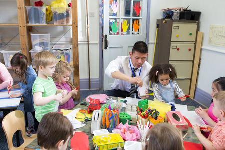 Male teacher with a class full of nursery students. They are all sat at tables using arts and crafts.の写真素材
