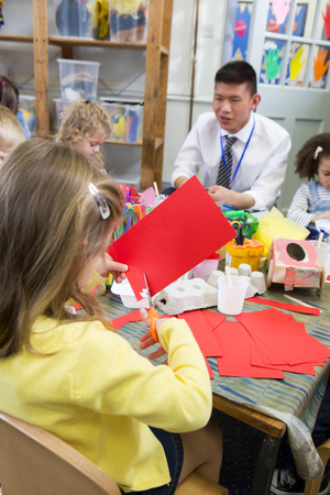 Little girl using scissors to cut shapes from a red piece of paper. She is in a nursery class with other students and a teacher.の写真素材