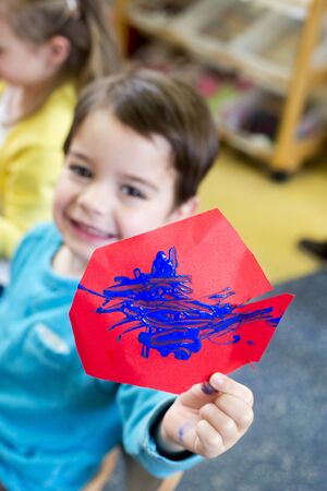 Little boy proudly holding up his painting to the camera. He has put blue paint on some red paper.の写真素材