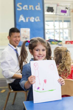 Little girl showing her drawing to the camera. She is in nursery and her teacher can be seen in the background.の写真素材