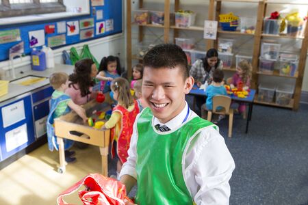 Teacher in a classroom. He is holding some aprons and smiling at the camera. The children are...