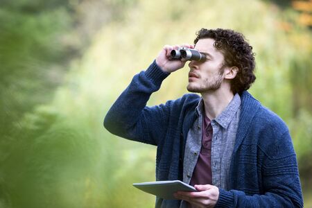 Young man analysing nature with binoculars. He is using a digital tablet to research.の写真素材