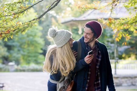 Young couple having fun together outdoors. The man is holding a smartphone while his girlfriend is jumping on his back. They are looking at eachother and laughing.の写真素材