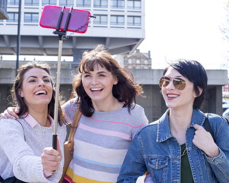 Three female friends taking a selfie in the city. They are using a smartphone on a selfie stick.の写真素材