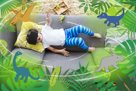 View from above of a little boy lying on his sofa with a box of dinosaurs. He is wearing a VR head set and there are dinosaur graphics around him.の写真素材