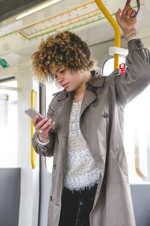 Woman standing on the train with a smart phone in her hand. She is holding the hand rail and looking...