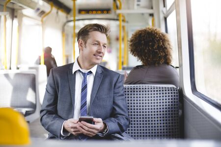 Formal businessman sitting on the train. He is holding a smartphone and looking out the window.の写真素材
