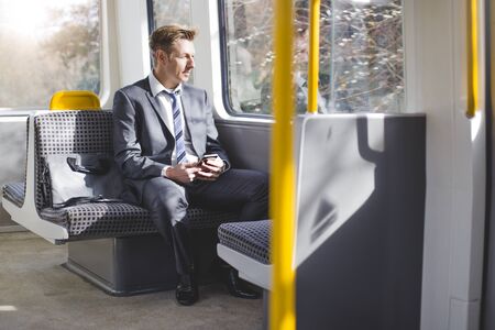 Formal business man sitting on the train with his bag. He is looking out the window and has a smart phone in his hands.の写真素材