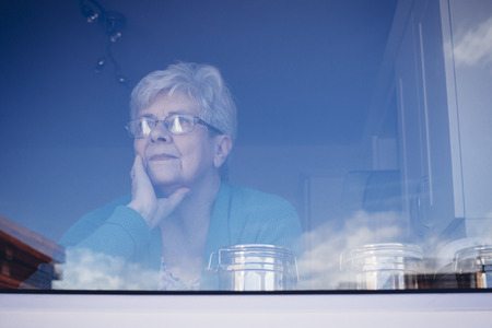 Senior woman looking out her kitchen window, deep in thought.の写真素材