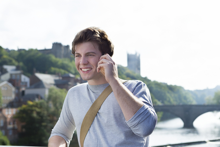 A young man uses his mobile phone outdoors next to a river. He is talking to someone. A cathederal can be seen in the background.の写真素材