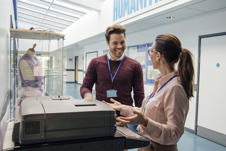 Two teachers are talking while standing at a printer in the school hall. 