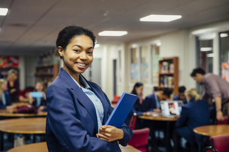 Teen student is posing for the camera in her classroom. She is holding a book and is wearing school uniform. の写真素材