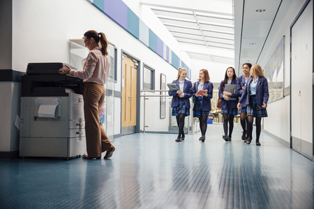 Group of teenage girls are walking down the school hall with books and laptops in their arms. They are talking and laughing as they walk and there is a female teacher using the printer.の写真素材