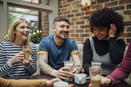 Friends are socialising over coffee in a coffee shop. They are laughing and talking with their drinks in their hands.の写真素材