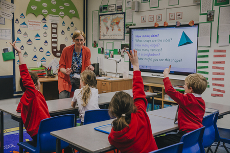 Female teacher is teaching shapes to her primary school students. She is asking hem a question and some of the students have their hand in the air to answer.の写真素材