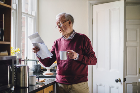 Senior man is standing in the kitchen of his home with bills in one hand and a cup of tea in the...