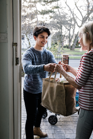 Teenage boy is delivering some groceries to an elderly woman. He is handing her a shopping bag at her front door.の写真素材