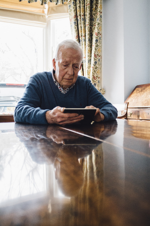 Senior man is sitting at his kitchen table in his home, looking sadly at a picture frame in his hands.の写真素材