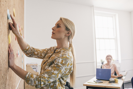 Female worker is pointing to a note on a cork board and is having a discussion with one of her coworkers, who is sitting at a desk in the background.の写真素材